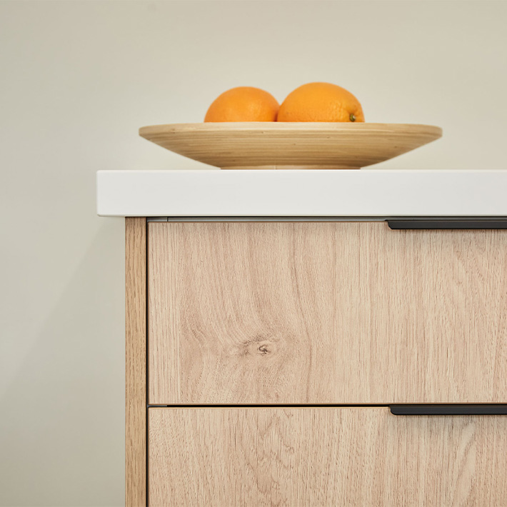 Two oranges in a wooden bowl on top of a modern cabinet with a white countertop.
