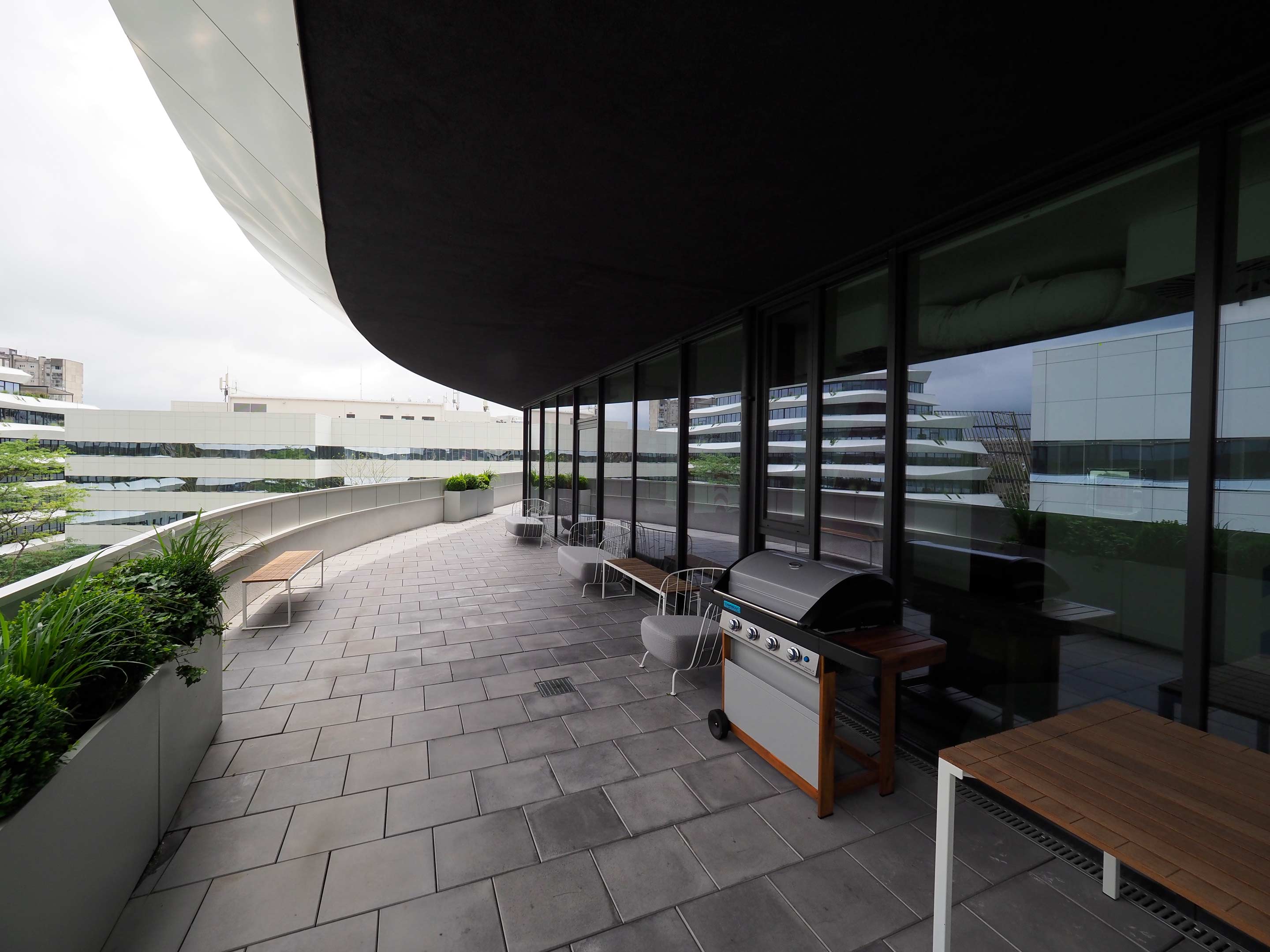 Modern terrace with a barbecue grill, seating, and planters under an overhanging roof with cityscape reflections.