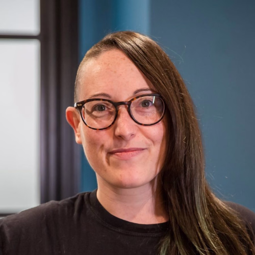 Natasha Hewlett company headshot with long hair wearing a black t-shirt, indoors with a blue wall in the background.