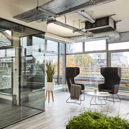 Modern office lounge area with large windows, black chairs, and exposed ceiling.