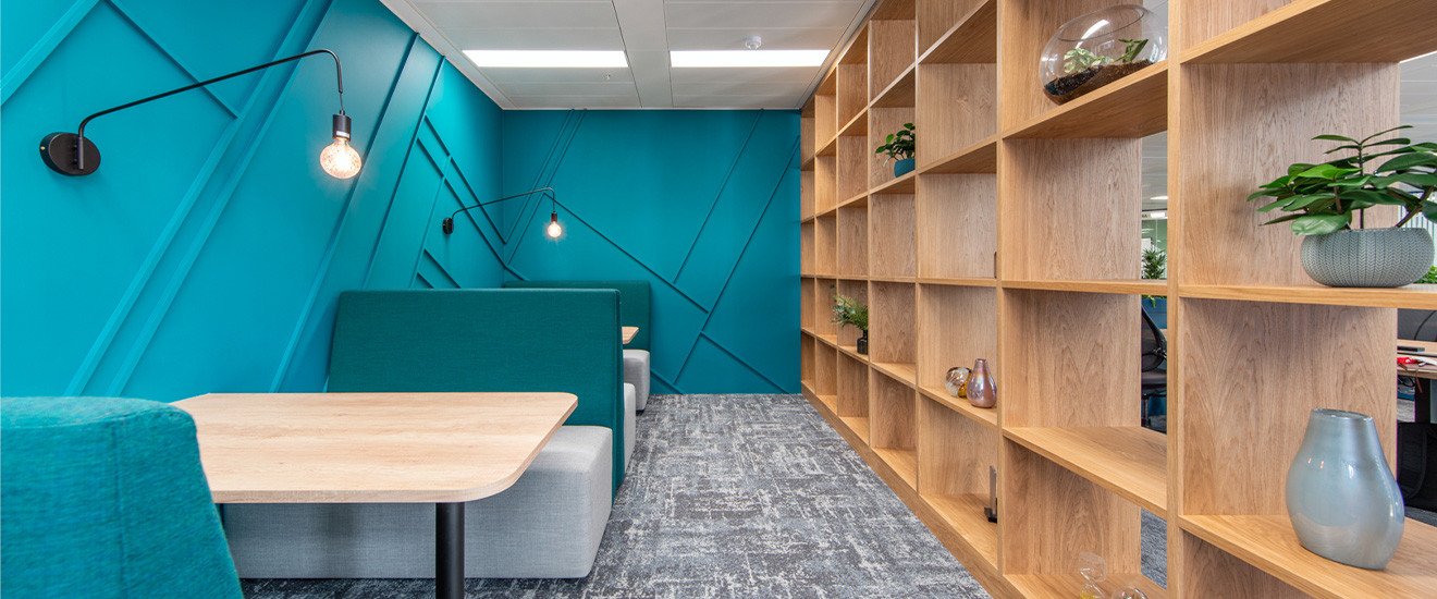 Modern office break area with teal walls, wooden bookshelves, a cozy booth with a green couch and a wooden table, and stylish hanging light fixtures.