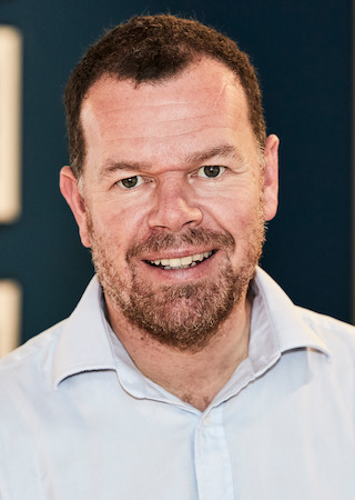 David Clowes employee headshot in a blue shirt against a blue backdrop.