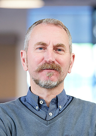 Phil Humphreys in a grey sweater over a blue patterned shirt against an indoor blurred background.