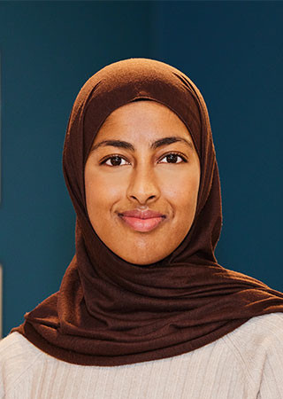 Rukshana Begum in a brown headscarf and cream sweater against a blue background.