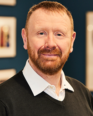 Sean Baxter company headshot in a black sweater over a white shirt, standing against blue office backdrop.