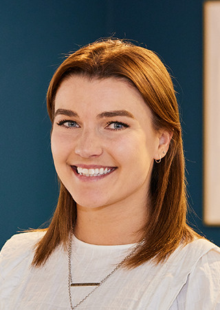 Company headshot of Steph Mcdermott wearing a white top, necklace and standing against blue backdrop.