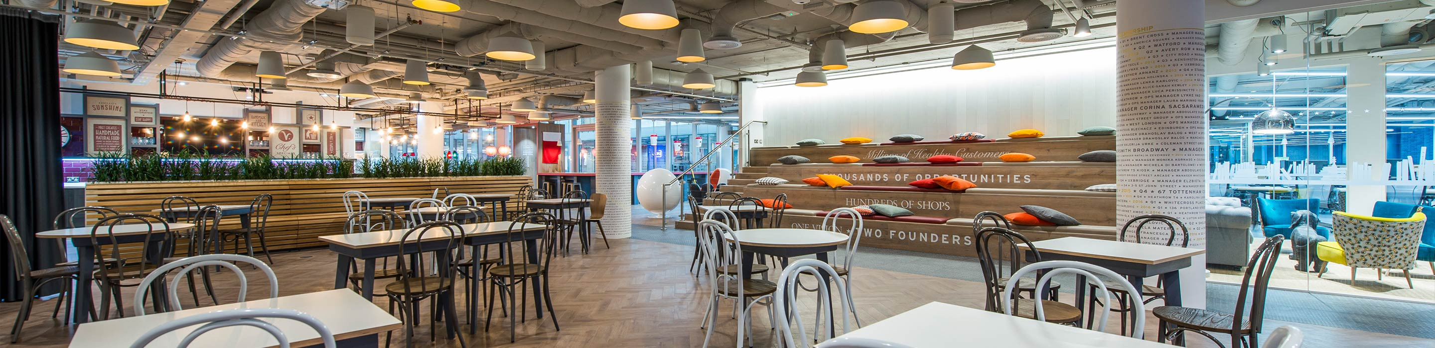 Modern cafeteria with varied seating options, decorative pillows on a bench, and inspirational wall murals.