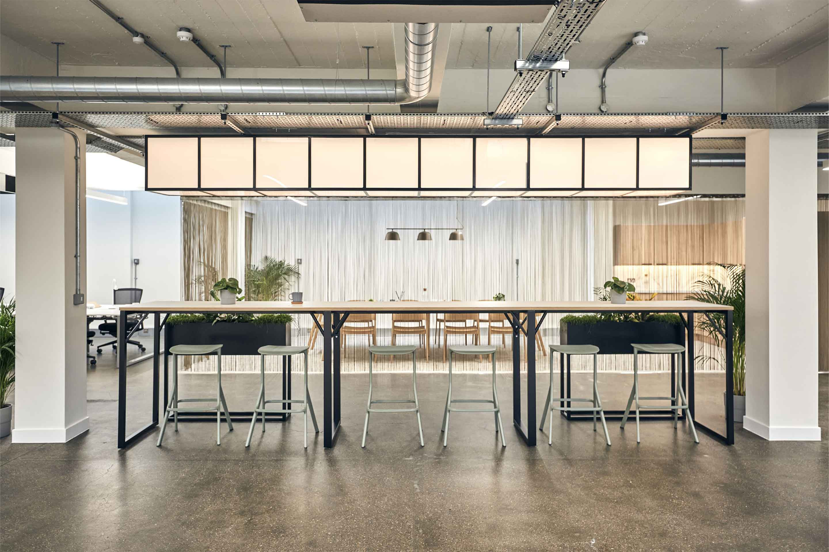 Modern office space with communal tables, hanging lights, and exposed ceiling.